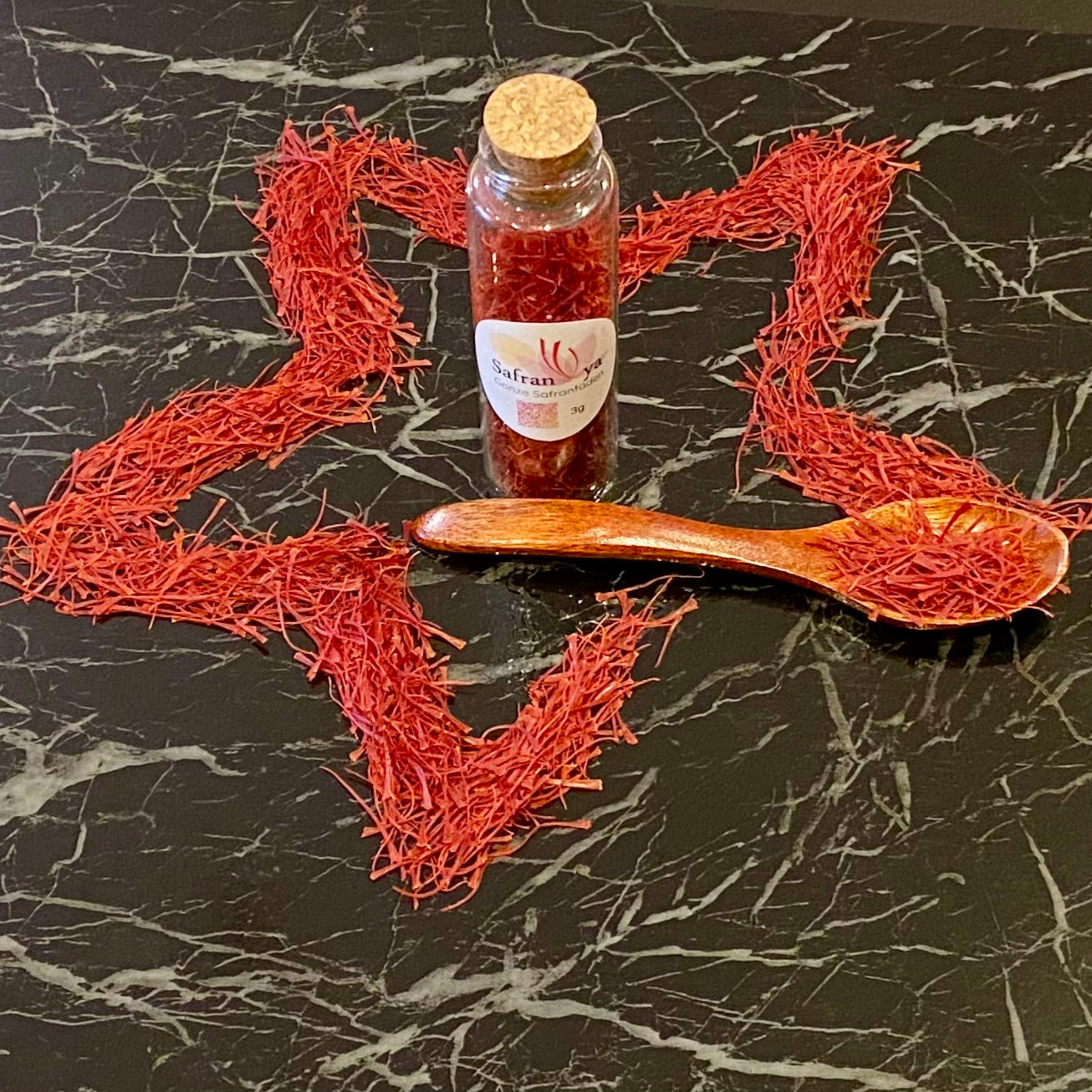 Safranfäden kunstvoll in Sternform arrangiert, mit Safranya Glasflasche und Holzlöffel auf schwarzem Marmortisch.