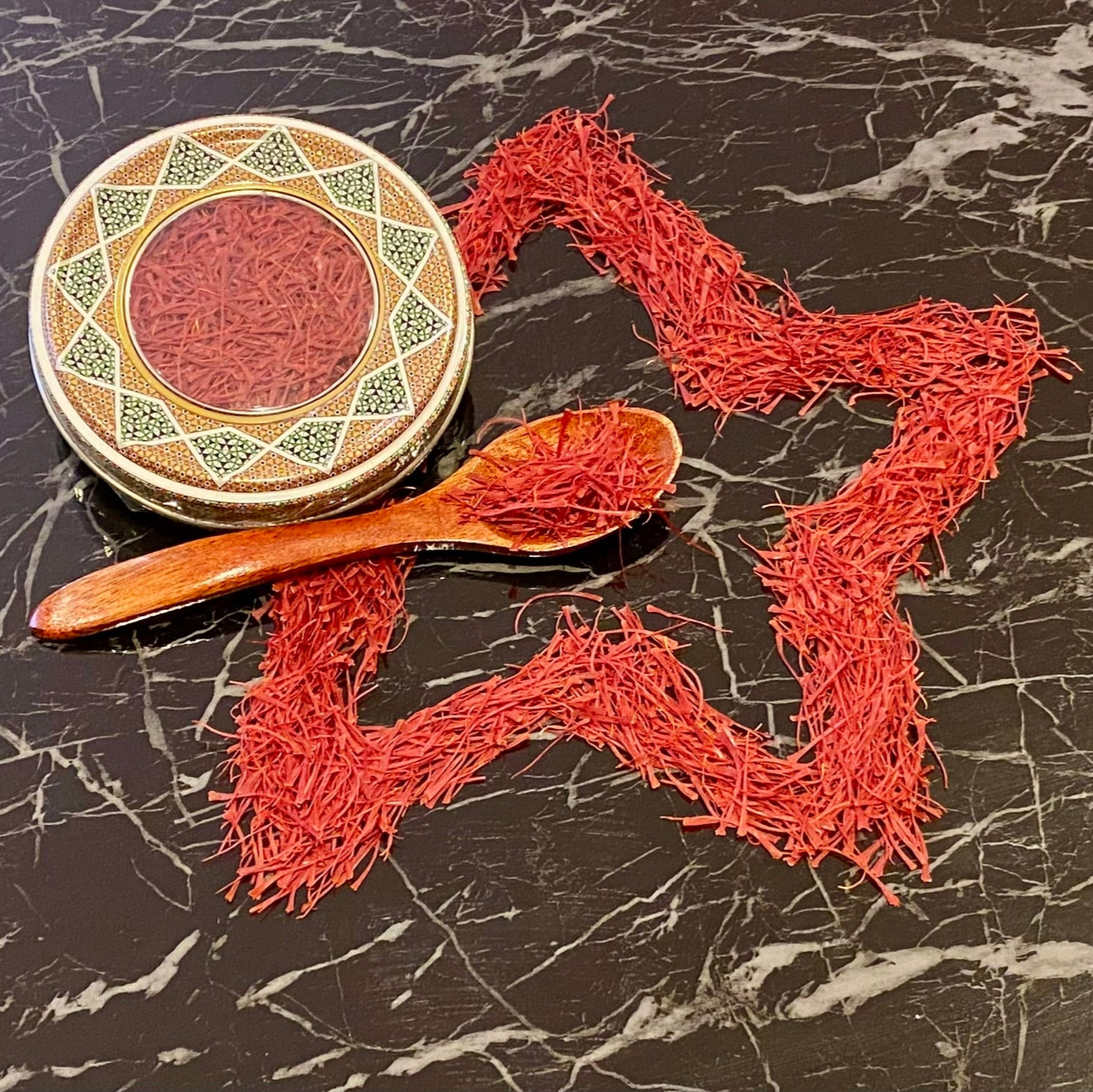 Hochwertige iranische Safranfäden in dekorativer Metalldose mit orientalischem Muster, Holzlöffel und kunstvoll zu einem Stern geformten Safranfäden auf schwarzem Marmortisch – Safranya Premium-Safran.