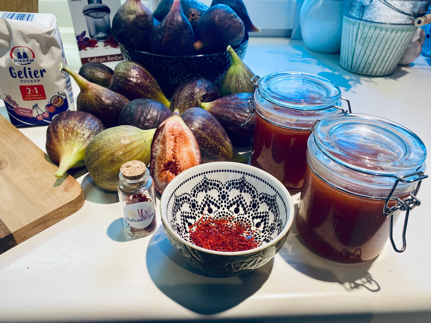 Ein Tisch ist mit frischen Feigen, einem Glas voller Safranfäden, einer Schale mit Safran, zwei Gläsern mit Feigenmarmelade und einem Paket Gelierzucker für die Zubereitung von Feigen-Safran-Marmelade.