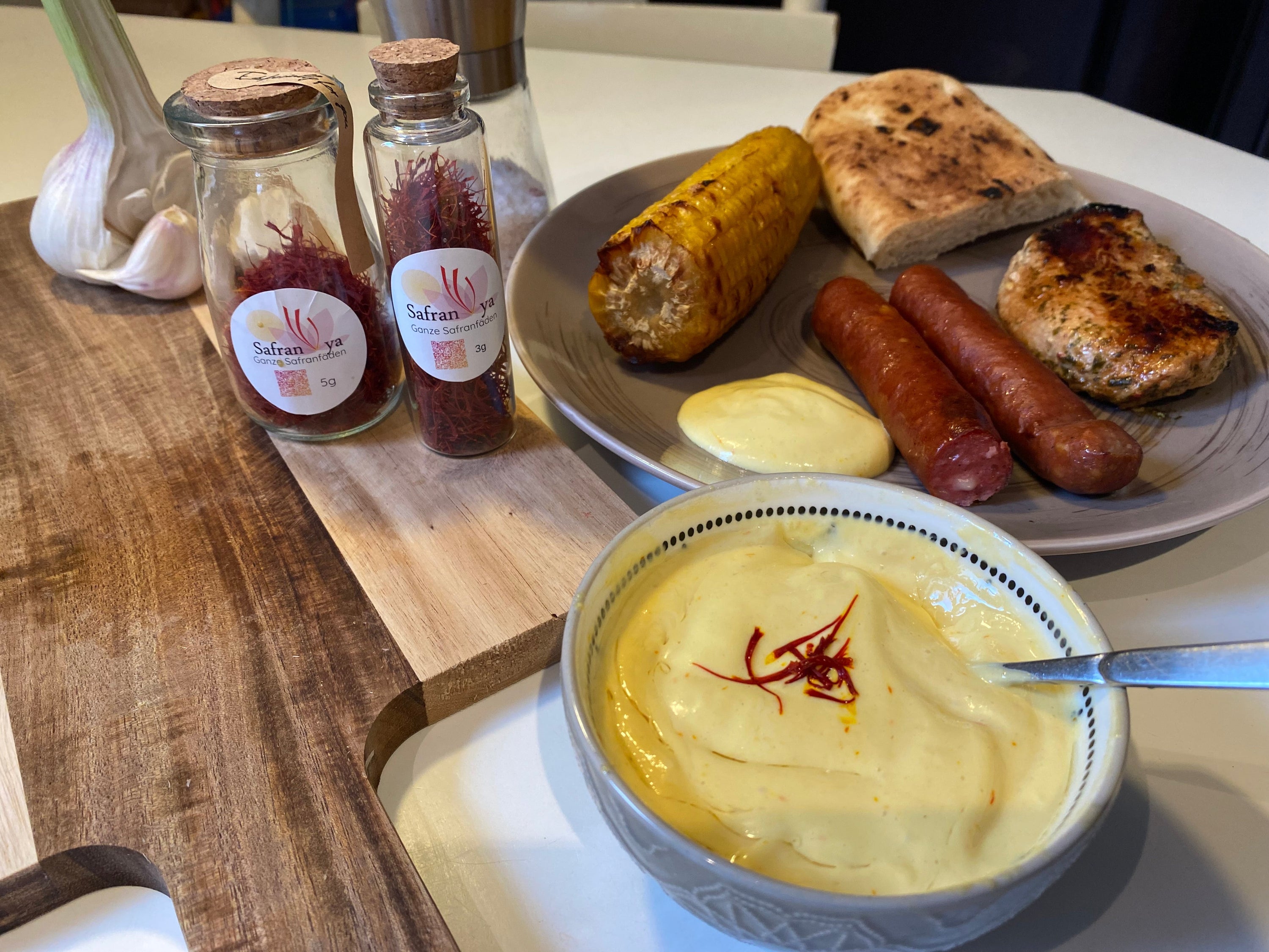 Ein Esstisch mit einer Platte, die Grillwürstchen, gegrilltes Hähnchen, Maiskolben und Fladenbrot enthält. Daneben stehen zwei Gläser mit Safran, eine Schüssel Safran-Dip und Knoblauch auf einem Holzbrett.