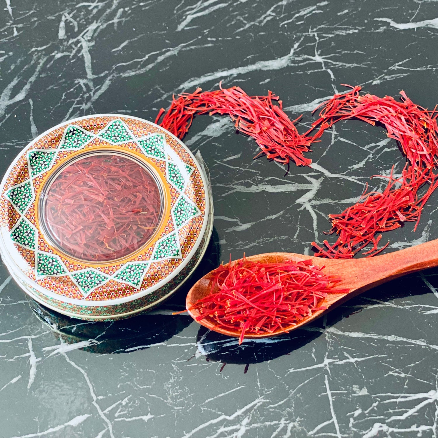 Orientalische Safran-Metallbox und Holzlöffel mit roten Safranfäden auf schwarzer Marmorplatte, daneben Safranfäden in Herzform gelegt.