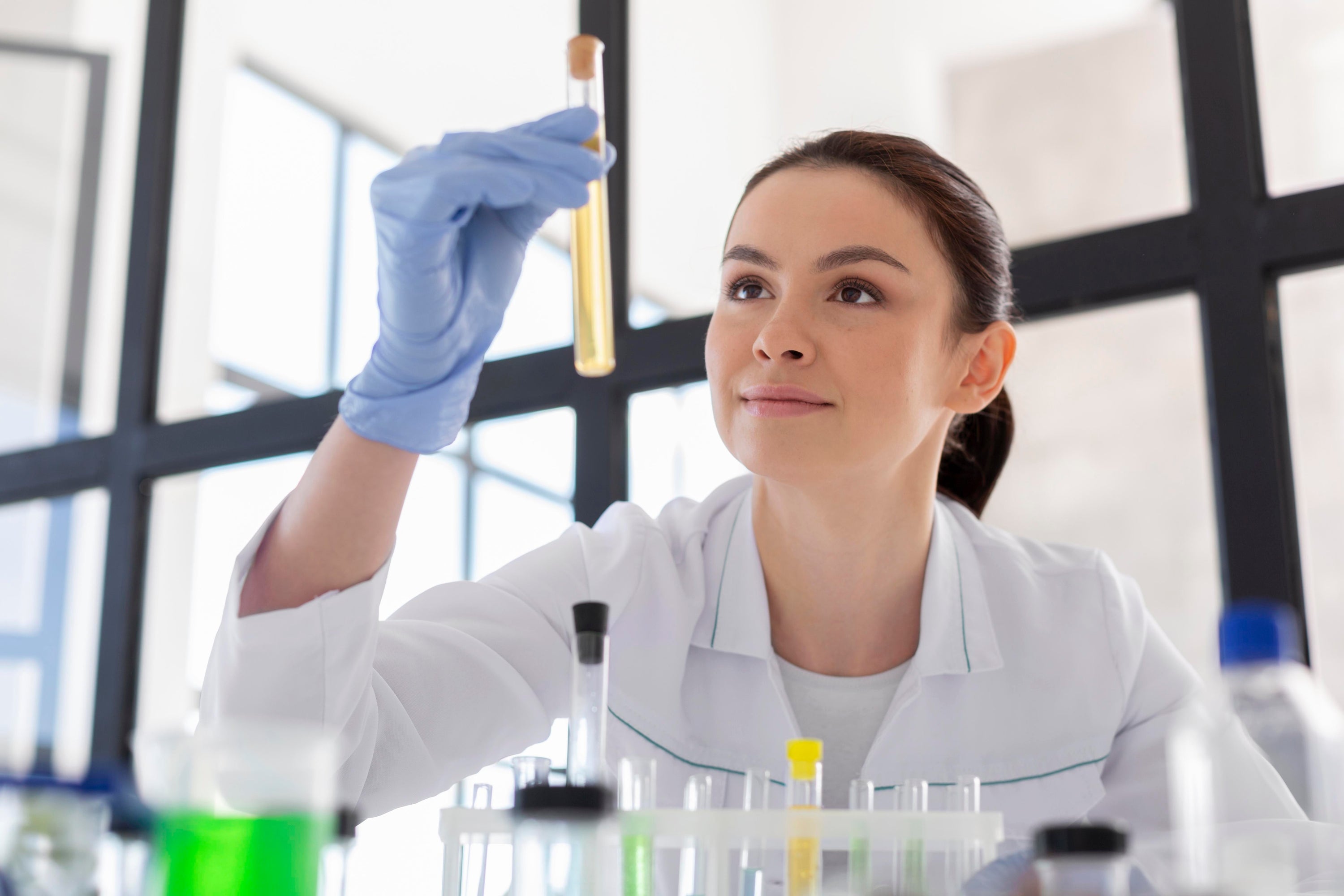 Junge Labormitarbeiterin im weißen Kittel und mit blauen Handschuhen, die lächelnd ein Reagenzglas mit gelber Flüssigkeit im Labor gegen das Licht hält.