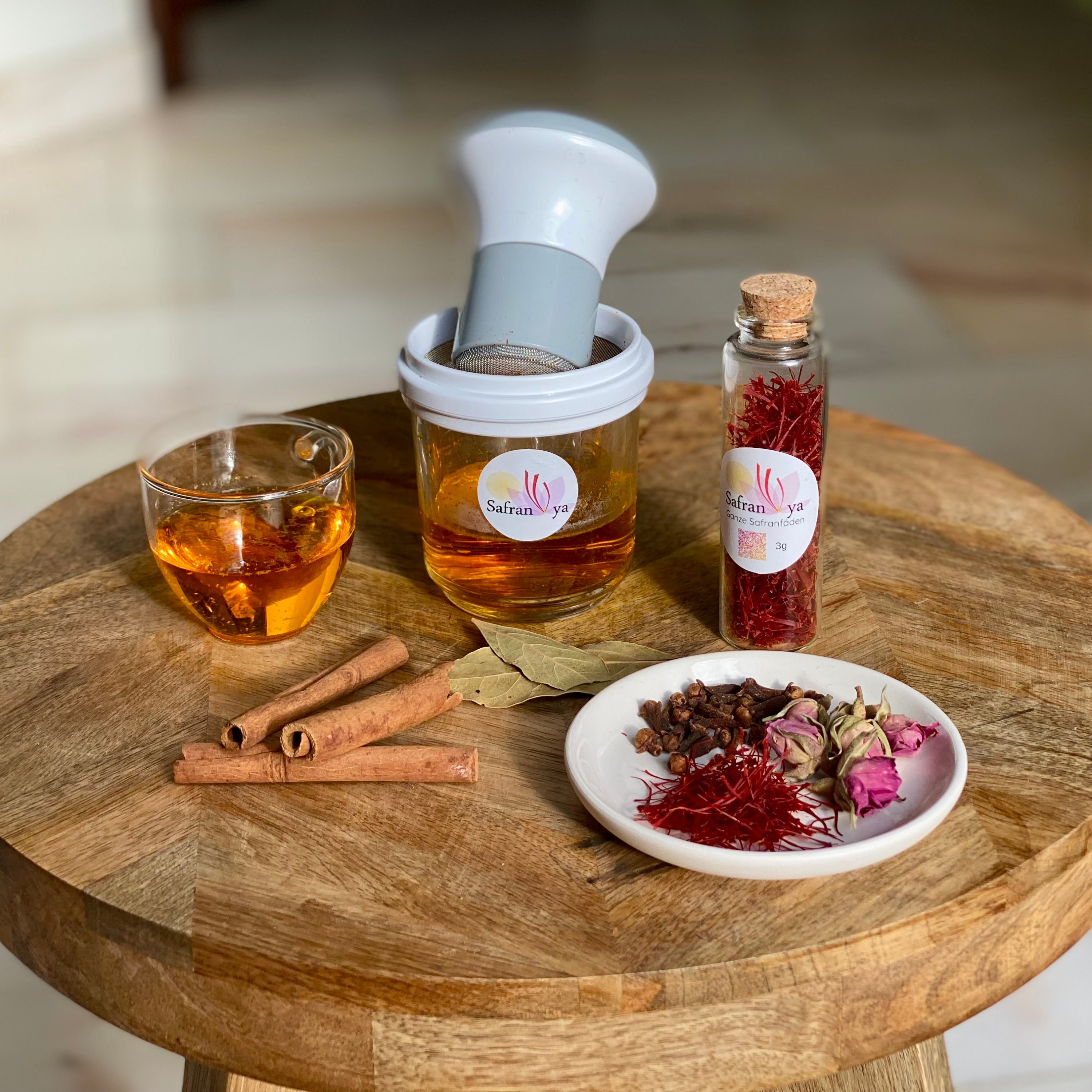 Safran Reibe von Safranya mit Glasbehälter, gefüllt mit warmem Safranwasser, daneben ein Glas Safrantee, Zimtstangen, Lorbeerblätter und eine Schale mit Safranfäden, Nelken und Rosenknospen auf einem Holztisch.