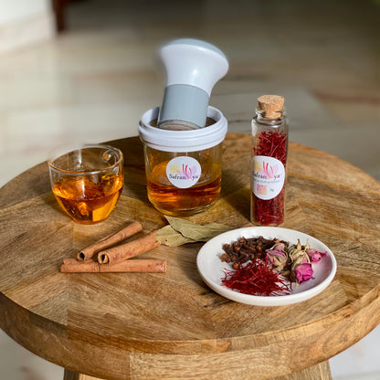 Safran Reibe von Safranya mit Glasbehälter, gefüllt mit warmem Safranwasser, daneben ein Glas Safrantee, Zimtstangen, Lorbeerblätter und eine Schale mit Safranfäden, Nelken und Rosenknospen auf einem Holztisch.