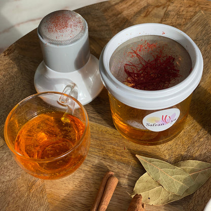 Detailaufnahme der Safran Reibe von Safranya: frisch gemahlene Safranfäden im Sieb, darunter ein Glas mit goldgelbem Safranwasser, daneben ein Glas Safrantee, Zimtstangen und Lorbeerblätter auf Holztisch.