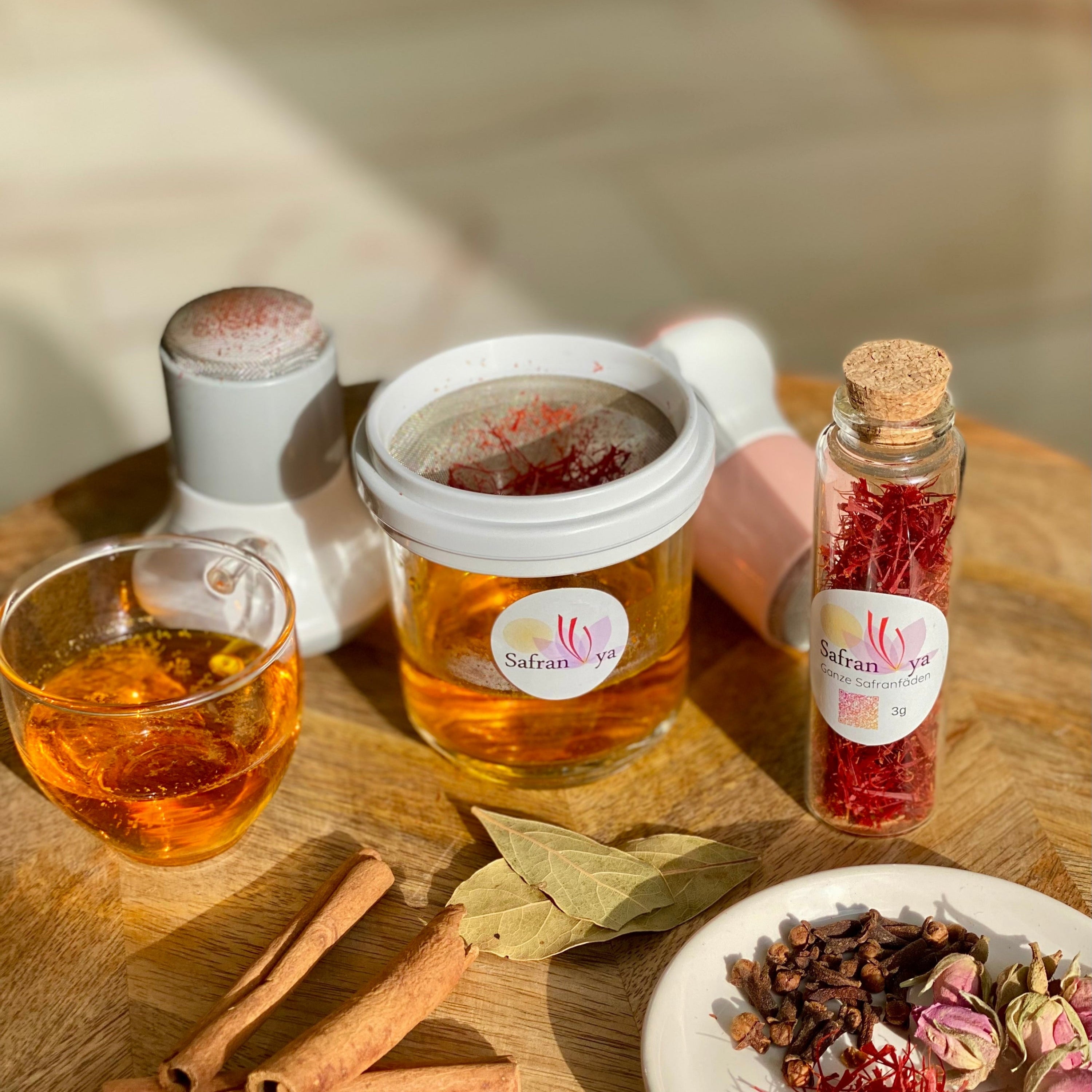 Safran Reibe im Einsatz mit Safranfäden im Sieb, daneben Safran im Glasröhrchen, ein Glas Safrantee, Zimtstangen, Lorbeerblätter und eine kleine Gewürzschale auf einem Holztisch.