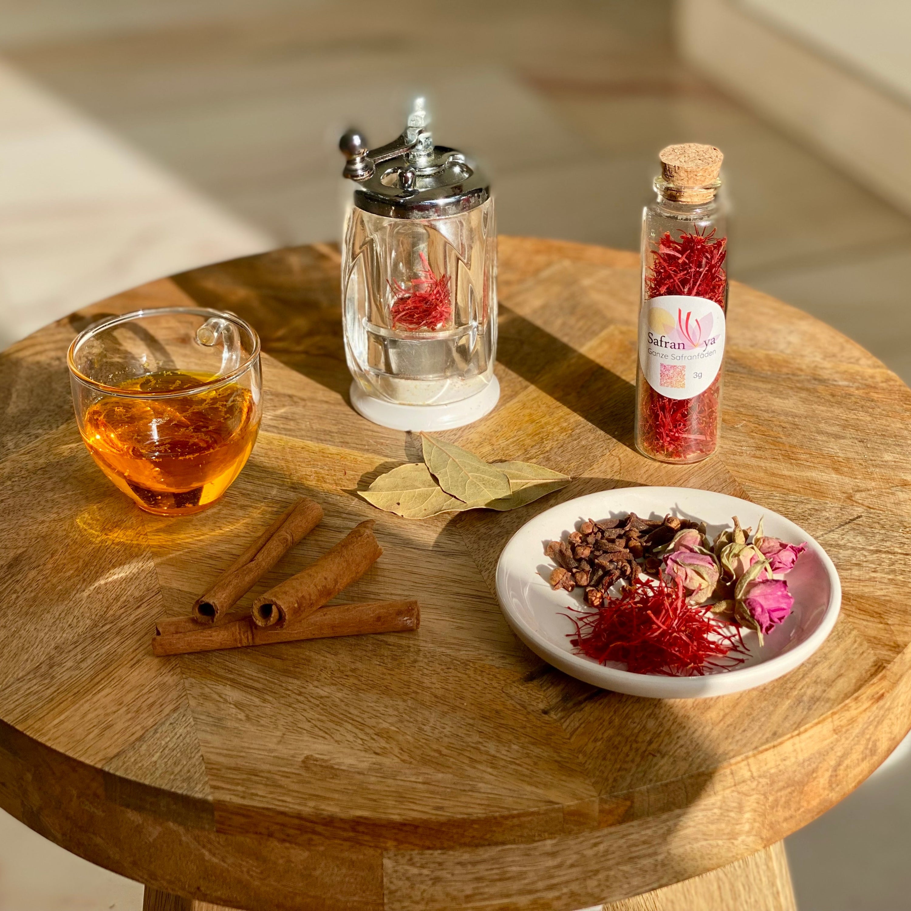 Glas Tee, Safranfäden in Glasröhre und Safranmühle auf Holztisch, umgeben von Zimtstangen, Lorbeerblättern, Nelken und Rosenknospen.