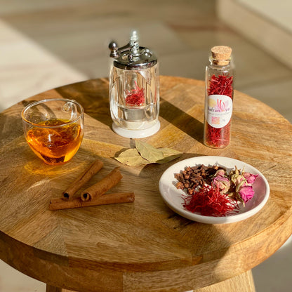 Glas Tee, Safranfäden in Glasröhre und Safranmühle auf Holztisch, umgeben von Zimtstangen, Lorbeerblättern, Nelken und Rosenknospen.