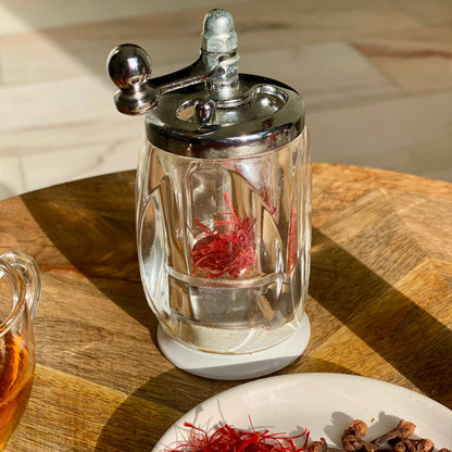 Nahaufnahme einer Safranmühle mit frischen Safranfäden, daneben ein Teller mit Nelken, Rosenknospen und Safran.