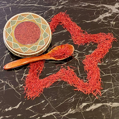 Hochwertige iranische Safranfäden in dekorativer Metalldose mit orientalischem Muster, Holzlöffel und kunstvoll zu einem Stern geformten Safranfäden auf schwarzem Marmortisch – Safranya Premium-Safran.